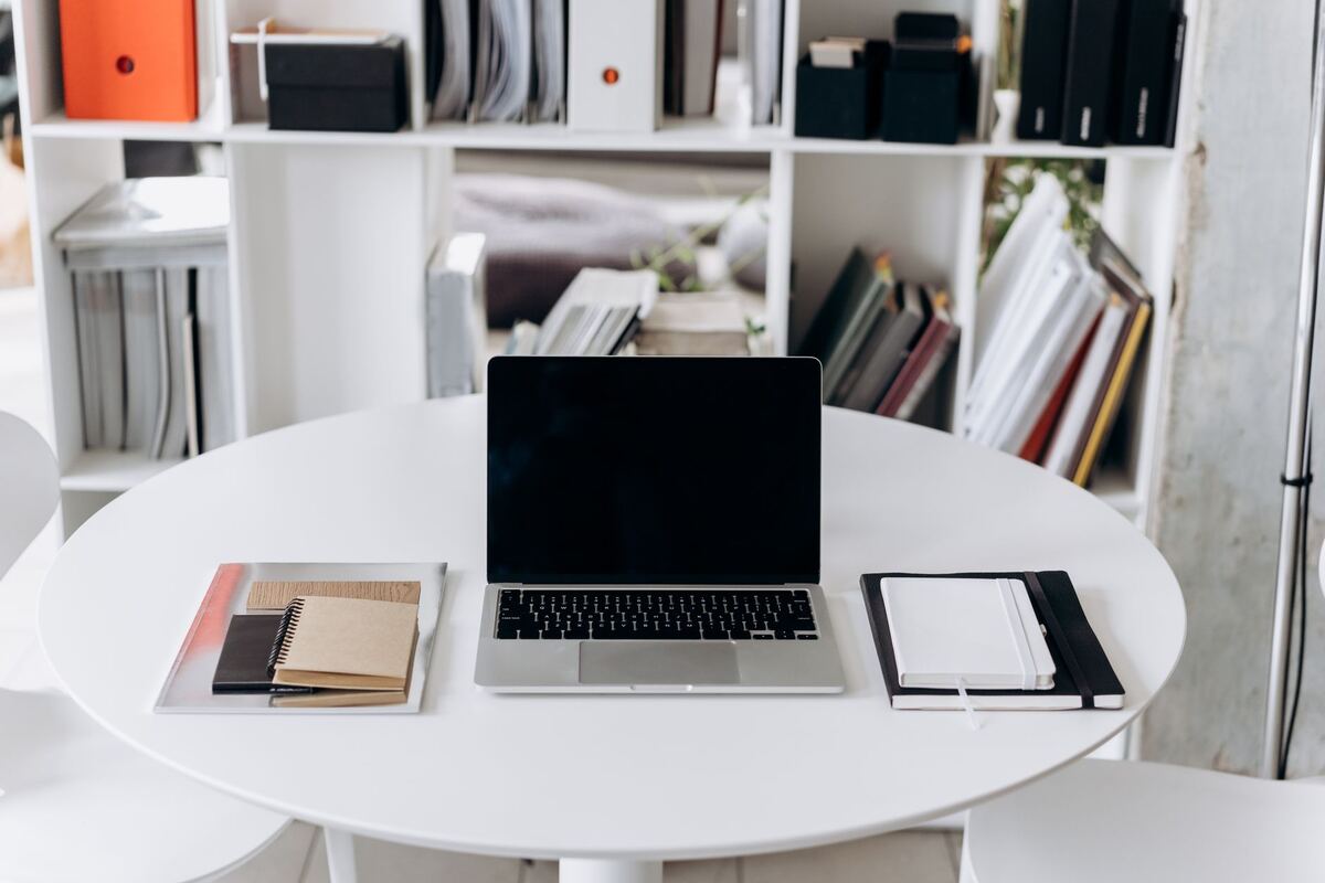 Laptop and notebooks on a round white table beside a bookshelf