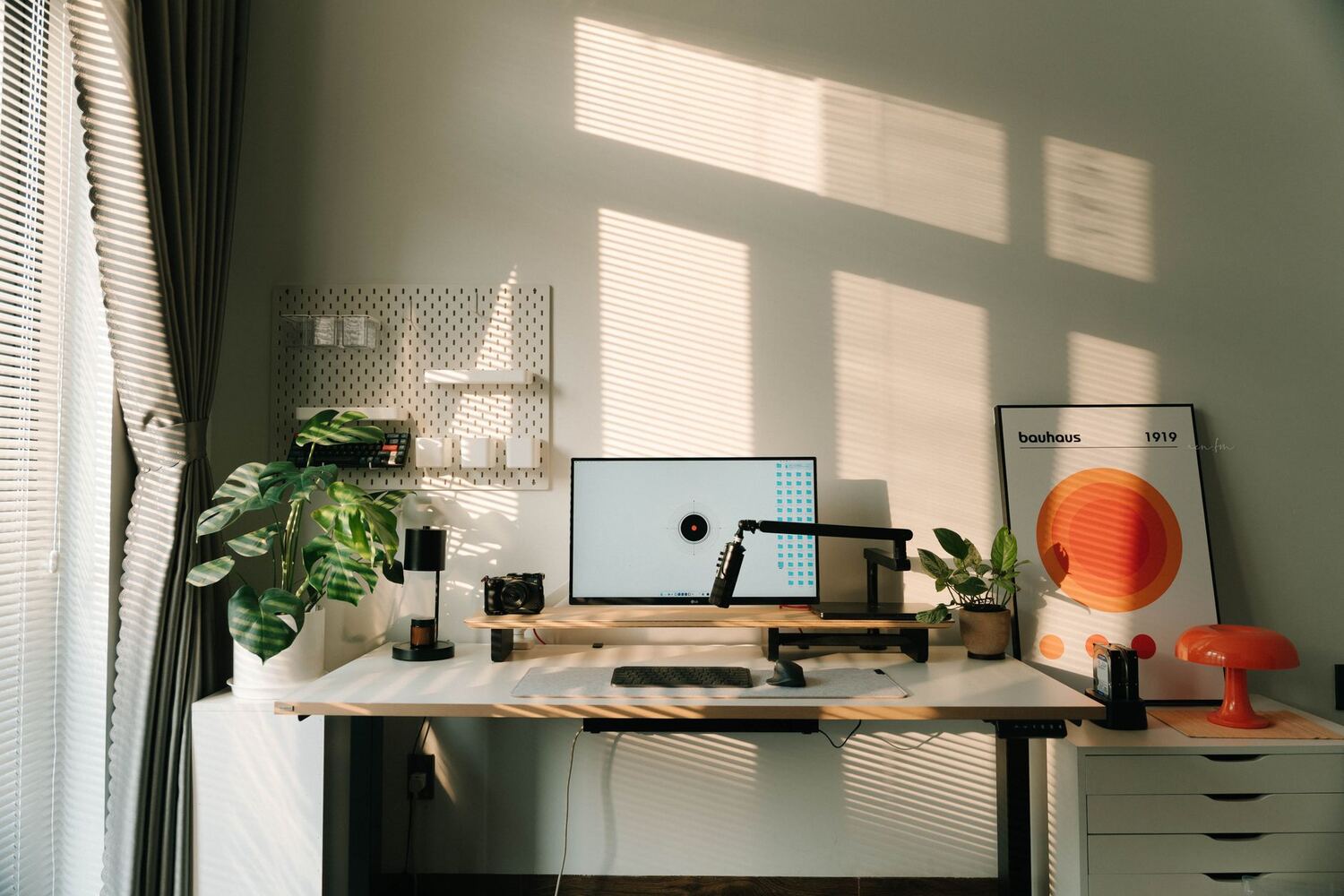Sunlit desk with a monitor, plants and a pegboard beside a shaded window